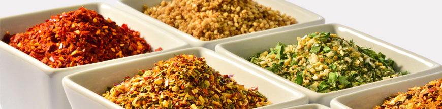 Six small white square bowls filled with various colorful dried spices and herbs, including red pepper flakes, coriander seeds, and chopped green herbs, arranged closely together on a white surface.