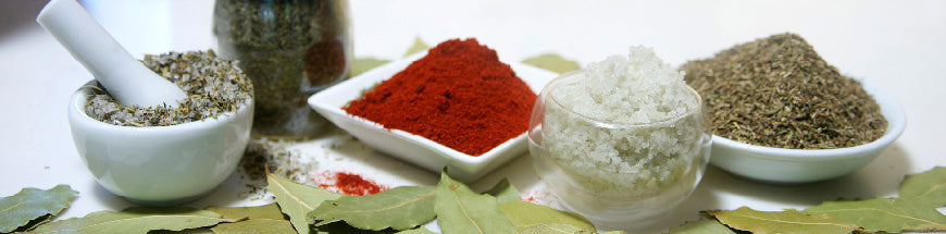 A variety of spices and herbs, including bay leaves, ground red pepper, salt, dried herbs, and a mortar and pestle, arranged on a white surface.