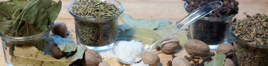 Small glass jars containing dried herbs, whole spices, and bay leaves are arranged on a wooden surface. A plastic spoon filled with salt and whole nutmegs are also visible.