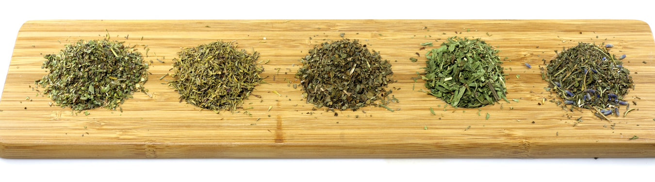 Five small piles of dried herbs arranged in a row on a wooden cutting board, each pile consisting of finely chopped green leaves and stems.