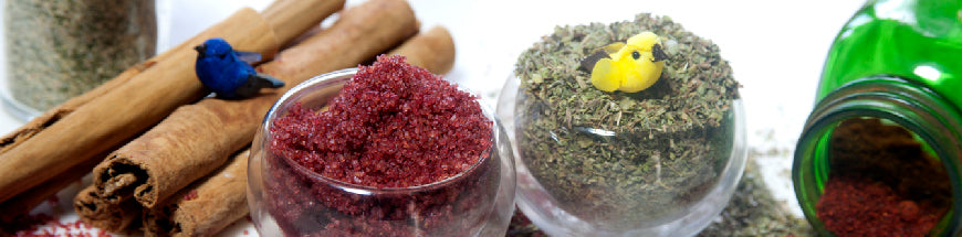 Spices in glass jars and glasses, including cinnamon sticks, red spice, and dried herbs, with small decorative birds placed on top. A green jar is tipped over, spilling contents.