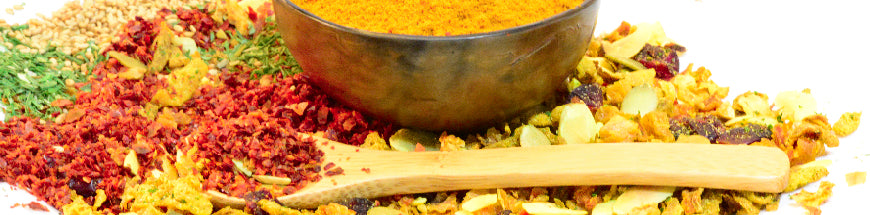 A variety of colorful dried spices and herbs are spread out on a surface, with a wooden spoon and a metal bowl filled with yellow spice powder in the background.