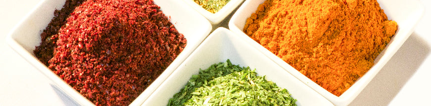 Four square white dishes filled with colorful ground spices and herbs—red, green, yellow, and a mix of greenish-yellow—arranged closely together on a white surface.