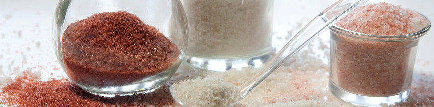Jars and a spoon filled with different varieties of salt, including white and pink coarse crystals, are arranged on a white surface with some salt scattered around.
