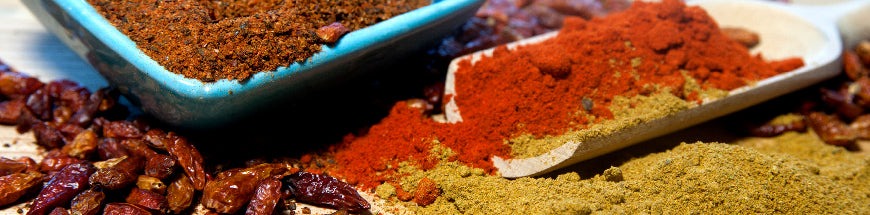 A close-up of various colorful spices, including ground red and green spices, dried chili peppers, and a bowl filled with a spice blend, arranged on a wooden surface.