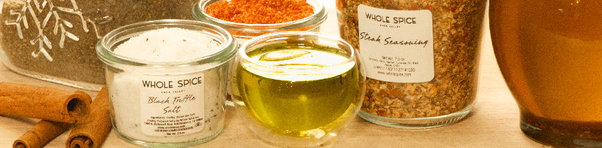 Various jars of spices, salt, and seasoning sit on a wooden surface next to cinnamon sticks and two small glass cups filled with olive oil. The scene is warmly lit.