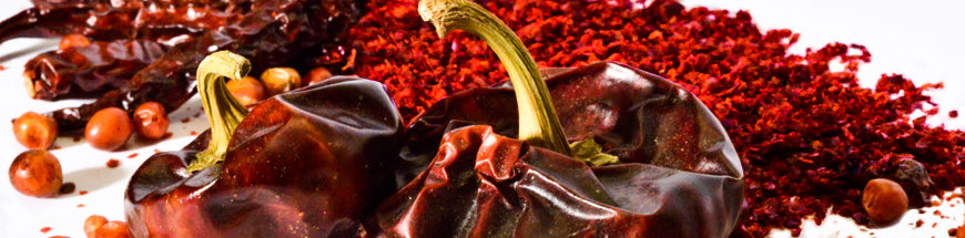 Close-up of dried red chili peppers, crushed red chili flakes, and scattered round brown seeds on a white surface.