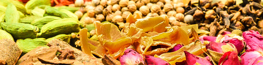 A vibrant assortment of dried spices and herbs, including green cardamom pods, dried rosebuds, bark, and various seed spices, arranged closely together.