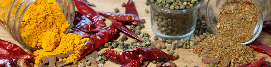 Spilled glass jars of turmeric powder, dried red chilies, green peppercorns, and a spice blend scattered on a wooden surface.