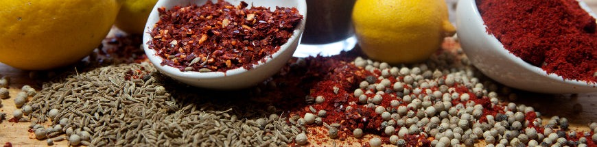 Close-up of various spices and seasonings, including cumin seeds, crushed red pepper, black pepper, white peppercorns, paprika, and whole lemons on a wooden surface.