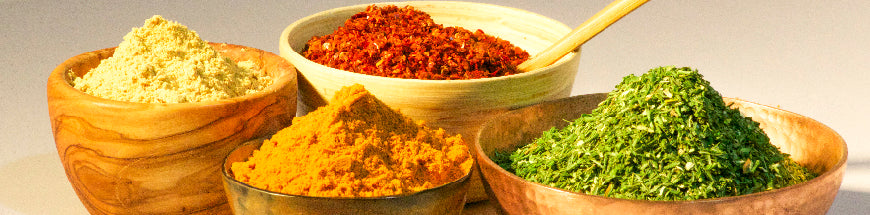Four wooden bowls filled with colorful ground spices and herbs, including yellow turmeric, green parsley, red chili flakes, and a light yellow powder, are arranged side by side on a light surface.