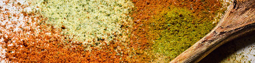 Colorful piles of ground spices in green, yellow, and orange shades are spread on a surface with a wooden spoon resting beside them.