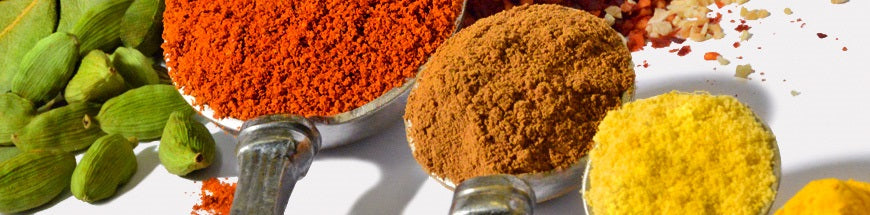 A close-up of various colorful spices including whole green cardamom pods, red chili powder, brown cinnamon, and yellow turmeric, arranged in scoops on a white surface.