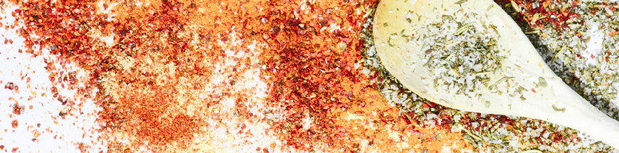 A mix of colorful ground spices, including red and green herbs, spread on a white surface with a wooden spoon filled with herb mix on the right.