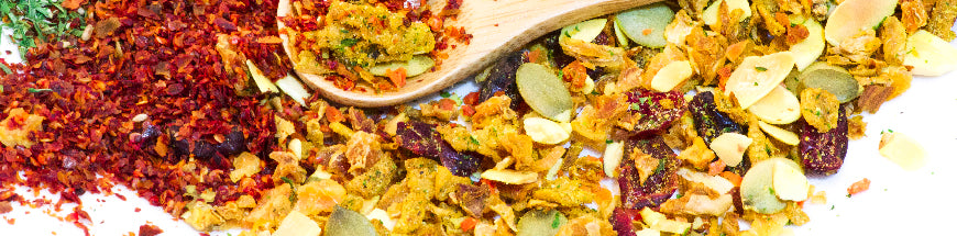 A close-up of colorful dried spices and fruit pieces, including red pepper flakes, yellow bits, seeds, and almond slivers, scattered on a white surface with a wooden spoon.