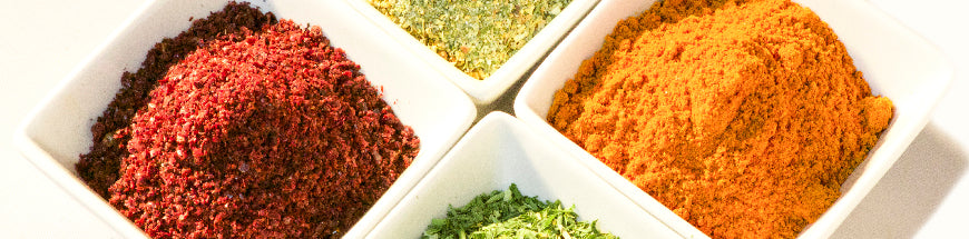 Four square white bowls filled with different colorful ground spices: red, yellow, green, and brown, arranged close together on a light surface.