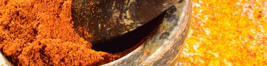 Close-up of a stone mortar and pestle grinding vibrant orange and yellow spices, with some ground spices scattered on a surface nearby.