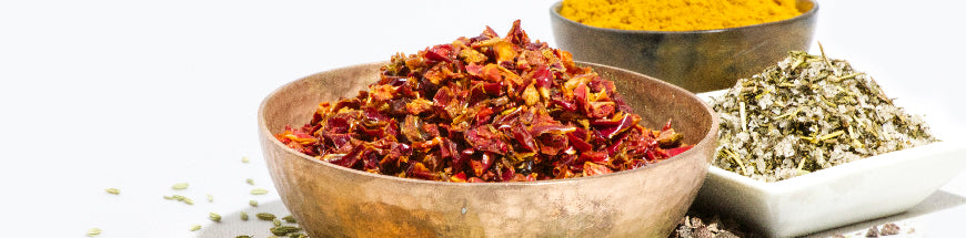 A copper bowl filled with dried red chili flakes, next to a square dish of dried herbs and a round bowl of yellow spice, all placed on a white surface.