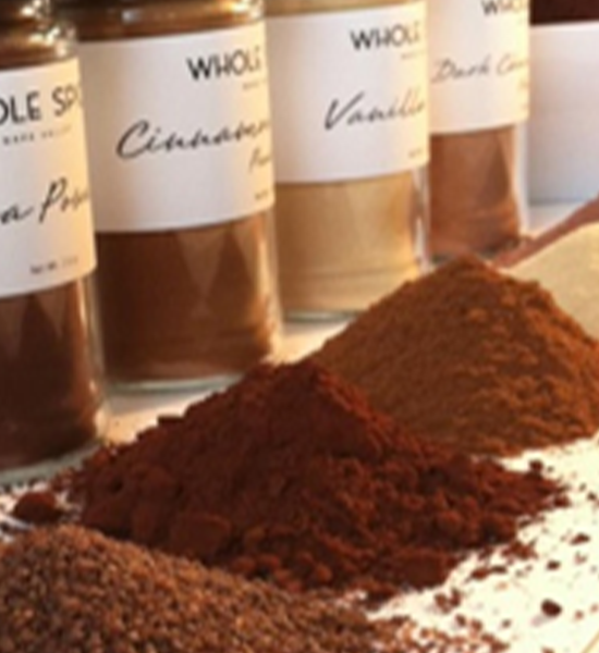 Several glass jars labeled with different spices, including cinnamon and vanilla, are in the background. In the foreground, small piles of powdered spices are spread out on a light surface.