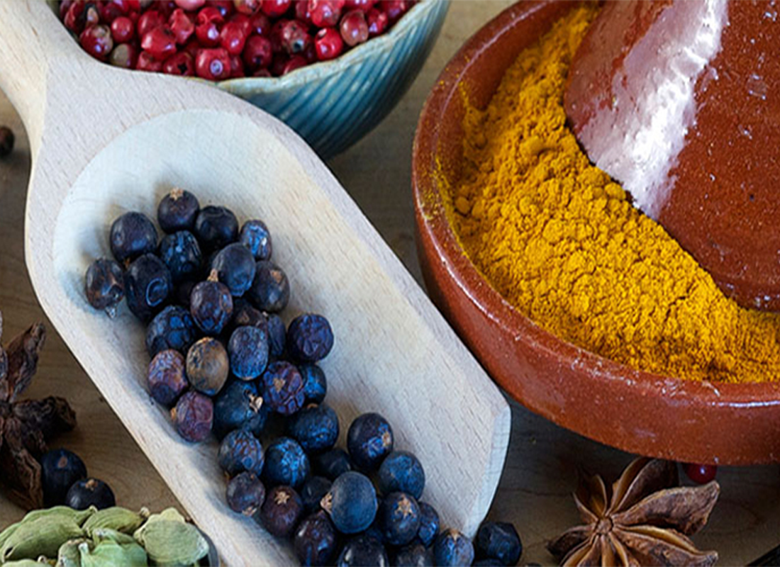 A wooden scoop filled with dried juniper berries sits next to a brown bowl containing bright yellow powdered turmeric and a blue bowl of red peppercorns; some cardamom pods and star anise are also visible.