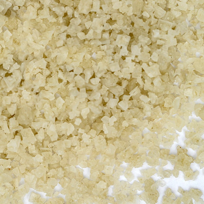 Close-up of many coarse French Gray Salt crystals from Whole Spice Inc., off-white to pale yellow, scattered across a white surface.