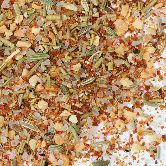A close-up of Whole Spice Inc.’s Napa Valley Rub reveals coarse salt, dried herbs, seeds, and red pepper flakes on a white surface—perfect as a seasoning rub or versatile marinade.