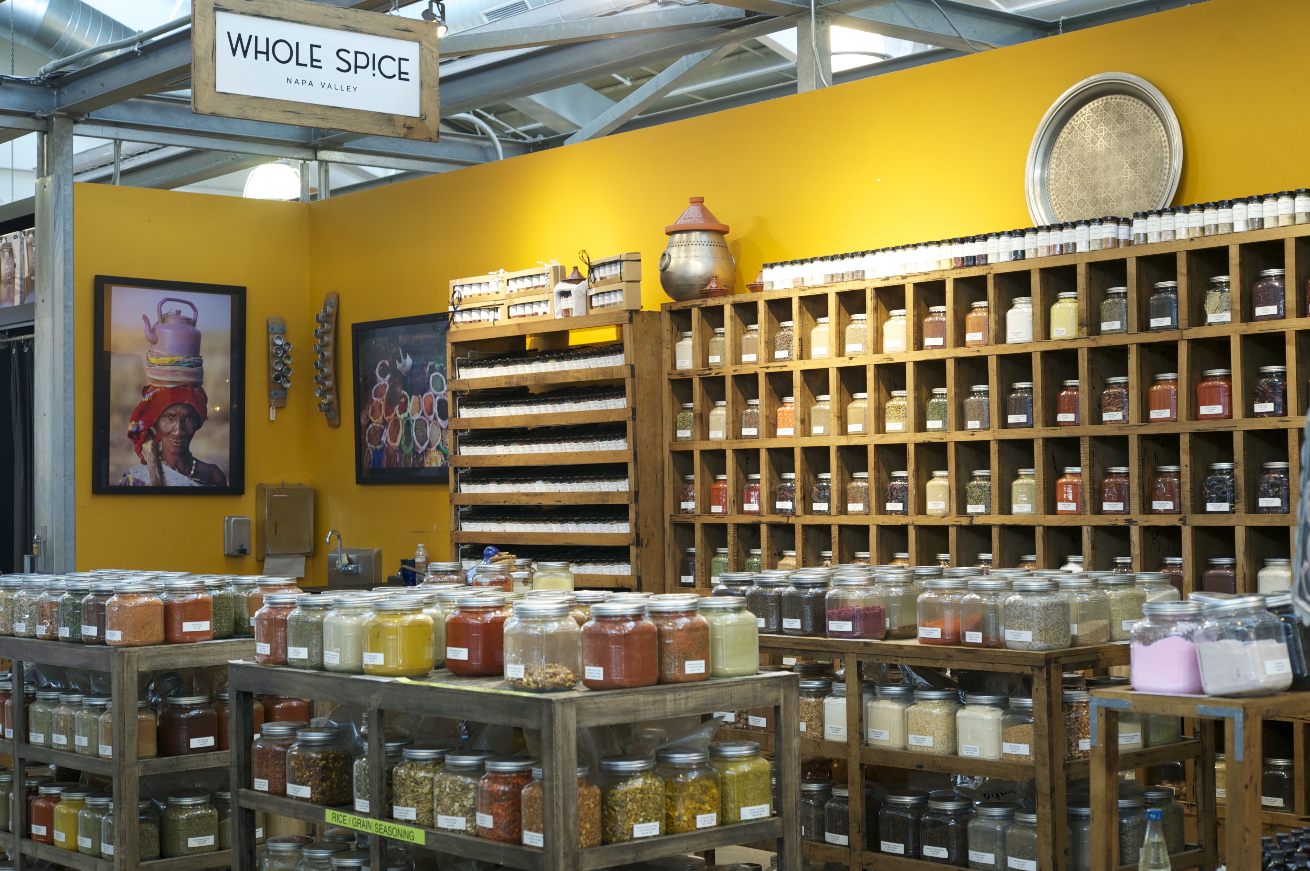 A colorful spice shop displays rows of glass jars filled with various spices on wooden shelves and tables. A yellow wall with art and a Whole Spice sign is visible in the background.