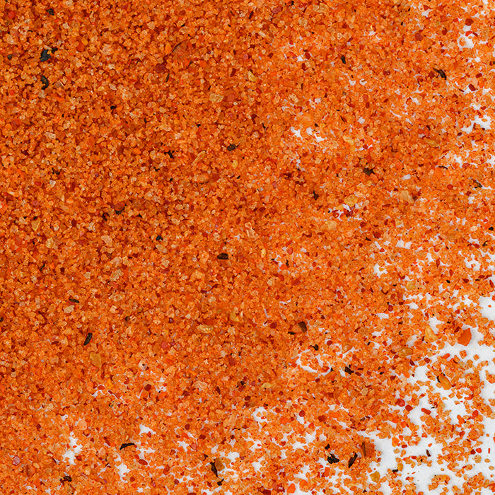 A close-up of Whole Spice Inc.'s Northwest Salmon Rub, a coarse orange-red seasoning blend for fish, is scattered on a white surface, revealing small colorful granules and specks in the gourmet mixture.