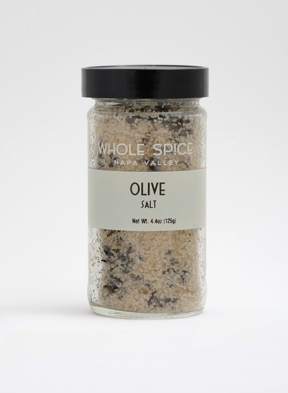 A clear glass jar with a black lid holds coarse Olive Salt by Whole Spice Inc., featuring visible dark olive pieces. Label states Net Wt. 4.4oz (125g), offering a gourmet sea salt experience.