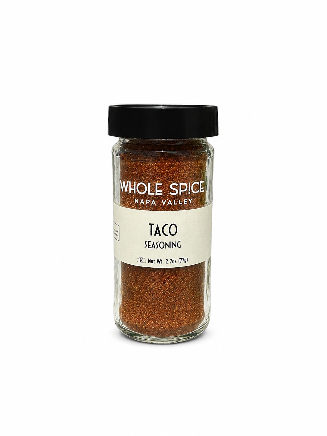A clear glass jar with a black lid labeled Whole Spice Inc. Taco Seasoning contains a reddish-brown Mexican seasoning blend. The net weight is 2.7 oz (77g), and the jar is displayed against a plain white background.