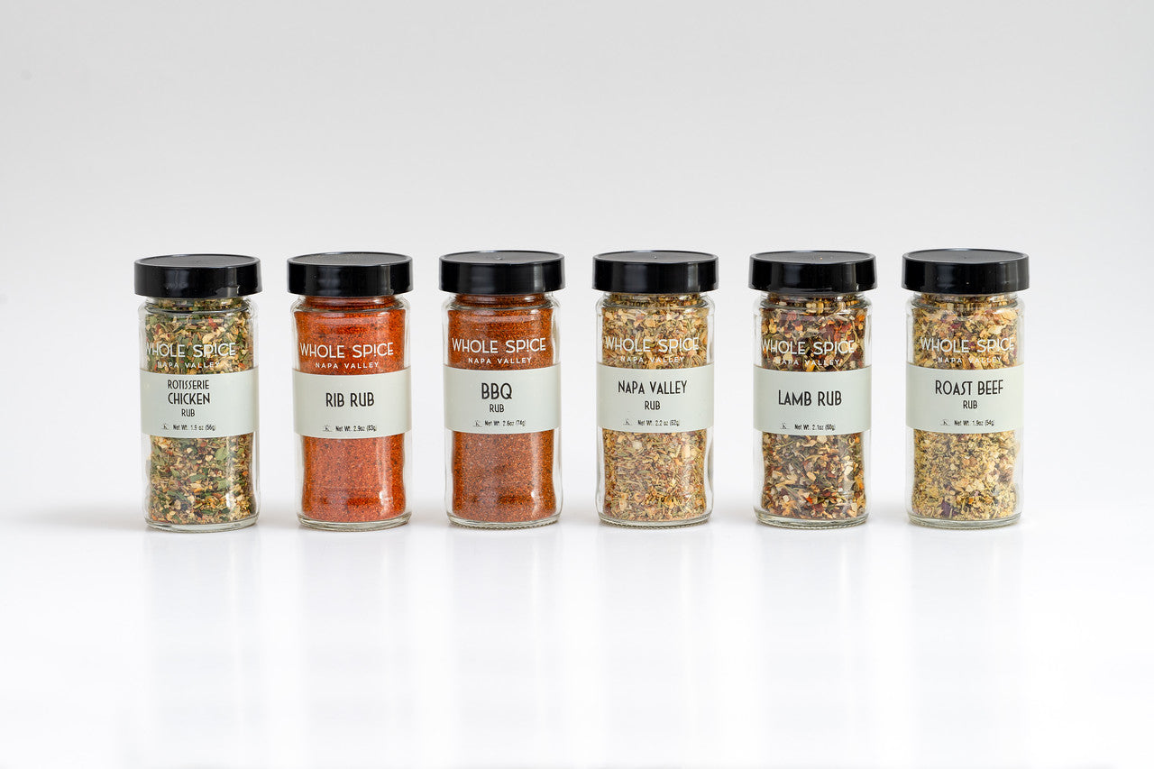 The Whole Spice Inc. Whole Spice Spice Rubs Gift Set includes six jars with black lids labeled Rotisserie Chicken, Rib Rub, BBQ, Napa Valley, Lamb Rub, and Roast Beef—essential spice rubs arranged in a handy set.