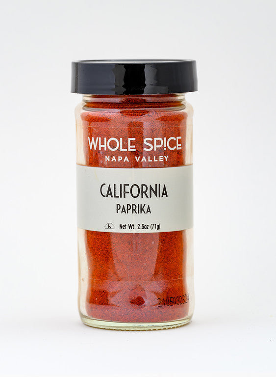 A glass jar of Whole Spice, Inc. California Paprika contains vibrant red paprika powder. The jar features a black lid, light gray label, and holds a net weight of 2.5 oz (71g).