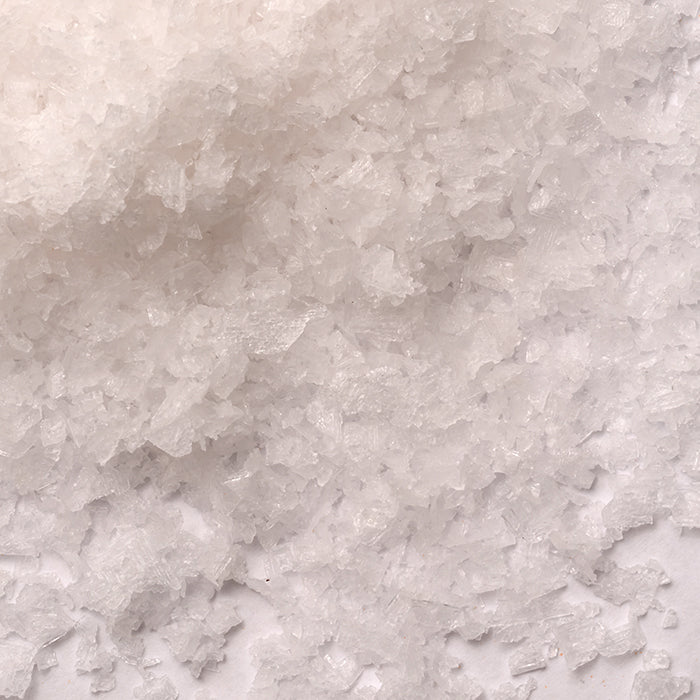Close-up of coarse, white English Sea Salt by Whole Spice Inc.—irregular, translucent crystals with a flaky texture and unique shapes scattered on a flat surface.