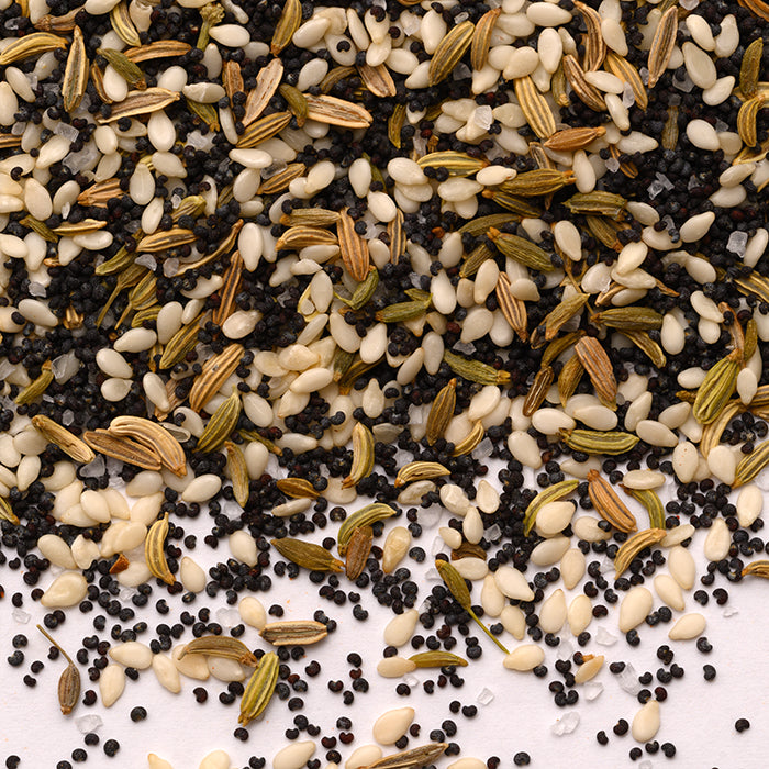 A close-up of Everything sans Bagel by Whole Spice Inc., showing a scattered mix of black mustard, white sesame, poppy, and greenish fennel seeds on a white surface.