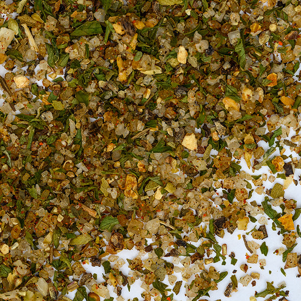 Close-up of Whole Spice Inc. Fish Seasoning—a vibrant mix of green, yellow, and brown herbs and spices—scattered on a white surface, ideal for adding all-purpose flavor to seafood dishes.