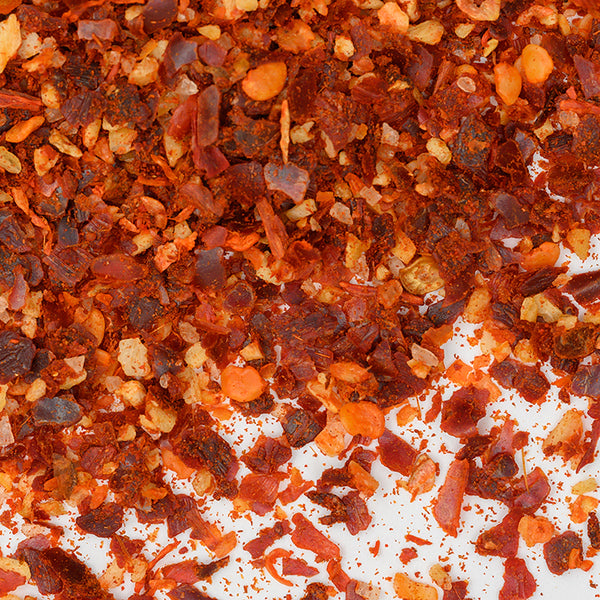 Close-up of Harissa Spice Coarse by Whole Spice Inc.—a vibrant blend of crushed red pepper flakes with red, orange, and yellowish pieces, seeds, and dried pepper skin scattered on a white surface.