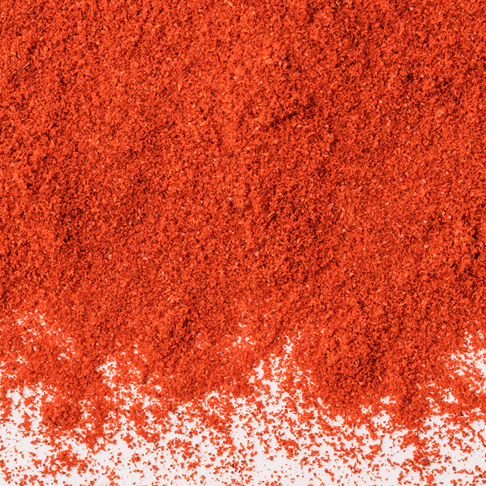 A close-up shows a heap of finely ground Hungarian Paprika by Whole Spice Inc., its vibrant red-orange powder spread unevenly over a white surface.