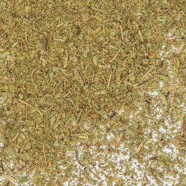 Close-up of Whole Spice Inc. Italian Seasoning Small Cut—a blend of dried, crushed herbs with a coarse texture, ideal for seasoning pasta or pizza. Scattered on a white surface, it features small pieces of leaves and stems.