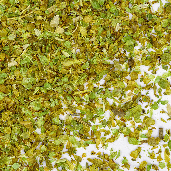 A close-up of Whole Spice Inc. Oregano Organic, a classic Mediterranean herb, shows dried leaves scattered on a white surface with visible small leaf fragments and pieces.