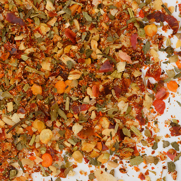 A close-up of Whole Spice Inc.'s Pizza Seasoning shows vibrant dried chili flakes, herbs, and seeds on a white surface—perfect for adding bold flavor to pasta sauces or using as a zesty topping on your favorite pizzas.