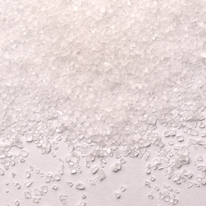 A close-up of coarse Pretzel Sea Salt crystals from Whole Spice Inc. scattered and piled on a plain white surface.