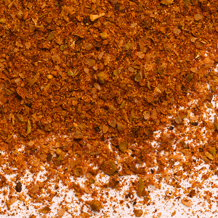 A close-up of Sazon by Whole Spice Inc., a reddish-brown Latin spice blend scattered on a white surface, highlighting its coarse texture with visible herbs and seeds—crafted as a traditional seasoning with no MSG.