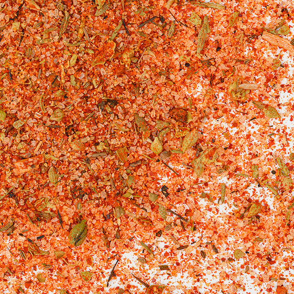 Close-up of Whole Spice Inc. Steak House Rub scattered on a white background, showing coarse pink salt crystals, red chili flakes, and green and brown dried herbs—ideal for seasoning beef.