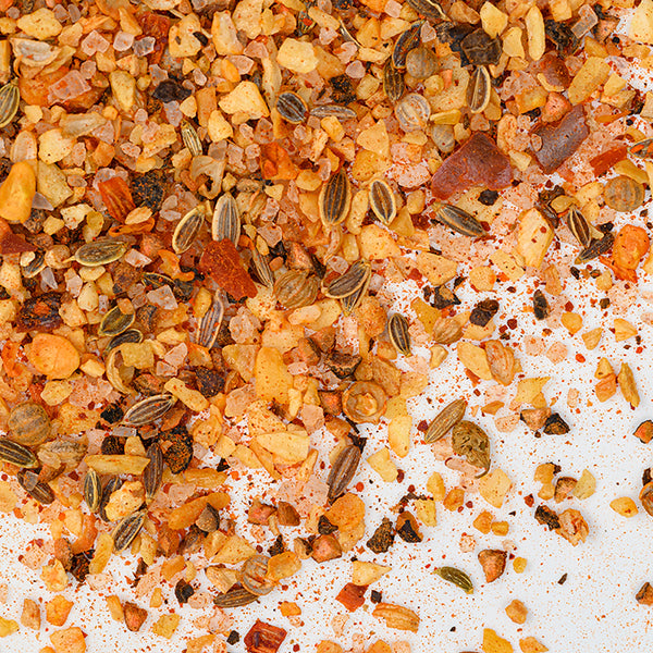 Close-up of Whole Spice Inc. Steak Seasoning showcasing colorful seeds, dried herbs, crushed peppers, and coarse salt sprinkled on a white surface.