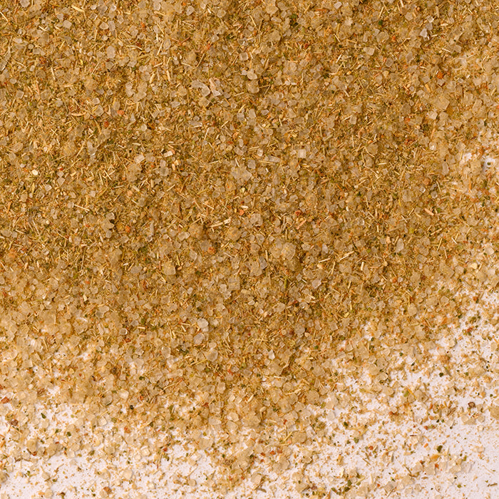 A close-up of Whole Spice Inc.’s Sweet Thai Seasoning shows coarse, light brown and greenish particles—an aromatic blend of Thai-inspired herbs, lemongrass, and salt—scattered unevenly on a white surface.