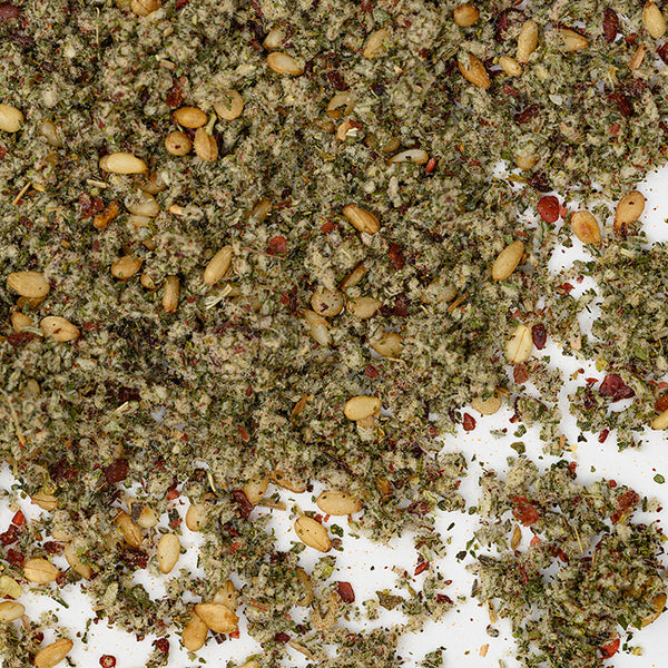 A close-up of Whole Spice Inc.'s Za'atar Spice Blend scattered on white shows its mix of dried herbs, sesame seeds, sumac, and pine nuts. The coarse texture and green-tan hues capture this authentic Middle Eastern seasoning.