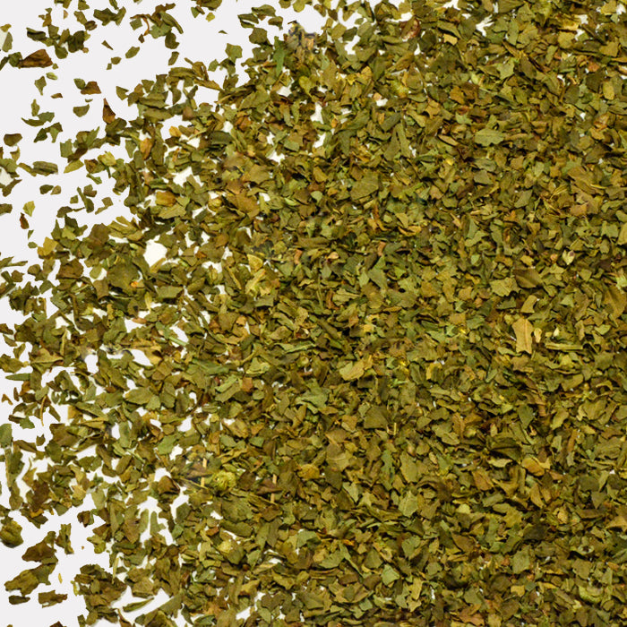 A close-up of Whole Spice Inc. Basil Organic, featuring dried green basil flakes scattered on a white background, some forming a pile and others spread loosely, showcasing their distinct texture.