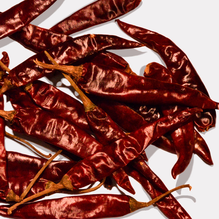 A pile of Whole Spice Inc. Puya Chili Whole peppers with shiny, wrinkled skin and stems, closely arranged on a light background, highlighting their signature fruity flavor.