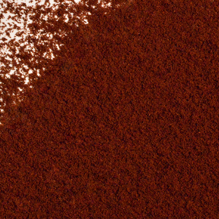 A close-up of a pile of finely ground Cloves Ground by Whole Spice Inc., a versatile spice often used in baking, spread across a white surface with some scattered granules in the upper left corner.