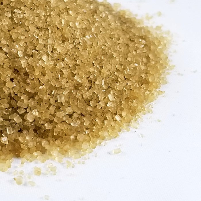 A close-up of Whole Spice Inc. Demerara Sugar granules, prized for their rich molasses flavor, scattered on a white surface.